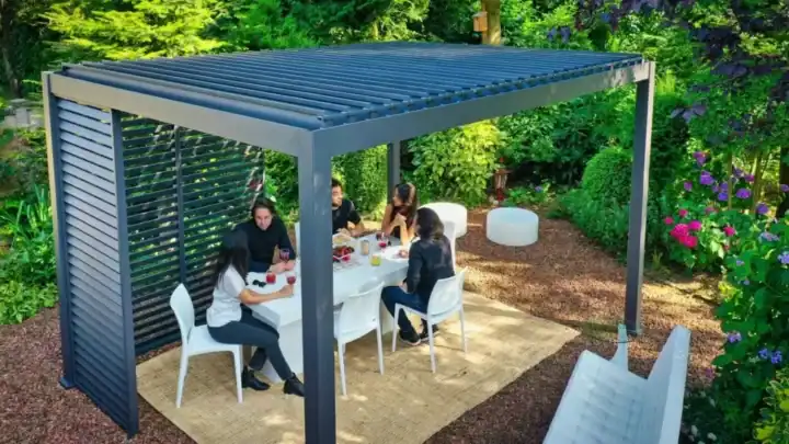 Menschen sitzen um einen weißen Tisch unter einer modernen Metallpergola in einem gepflegten Garten. Die Pergola hat ein Lamellendach und Seitenwände. Zu der Szene gehören weiße Stühle, Getränke, Snacks und Grünpflanzen mit farbenfrohen Hortensien. Unter dem Tisch liegt eine gewebte Tischdecke.