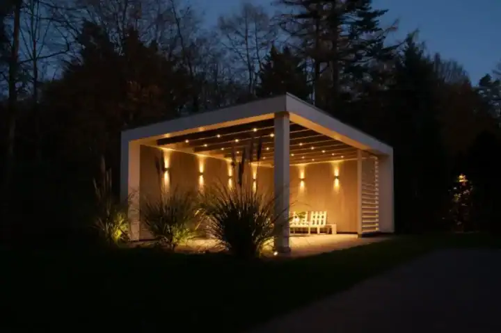 Ein moderner, beleuchteter Pavillon mit Flachdach in der Nacht, umgeben von hohem Gras und Bäumen. Im Deckenelement des Pavillons sind warme Lichter integriert, die einen gemütlichen Glanz verbreiten. Im Inneren der Struktur sind zwei weiße Stühle zu sehen.