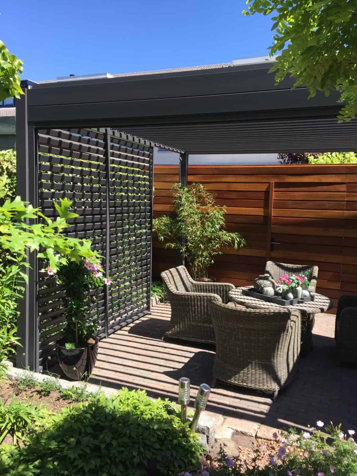 Außenpatio mit Rattanstühlen und einem Tisch unter einer Pergola. Sonnenlicht wirft Schatten durch das Lamellendach. Umgeben von Grün und einem Holzzaun bietet der Raum eine ruhige Kulisse vor strahlend blauem Himmel.