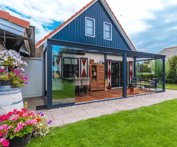 Ein modernes Haus mit dunkelblauer Außenfassade und einer rot überdachten Terrasse mit eleganten Glaswänden. Im Inneren gibt es einen gemütlichen Sitzbereich und eine Bar, geschmückt mit Topfblumen und eingerahmt von einem gepflegten Rasen im Vordergrund unter einem teilweise bewölkten Himmel.