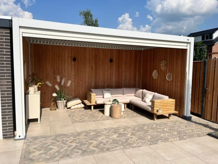 Eine moderne Außenterrasse verfügt über eine Holzumrandung und ein helles L-förmiges Sofa, ergänzt durch Kissen und kleine Pflanzen. Ein kleiner Tisch und gewebte Wanddekorationen verstärken das gemütliche Ambiente unter einem teilweise bewölkten Himmel, alles unter dem Schutz einer stilvollen Lamellenüberdachung.