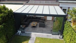 Ein moderner Outdoor-Sitzbereich mit Lamellendach manuell, Glaswänden, einem grauen Ecksofa und einem Couchtisch auf grauen Fliesen. Umgeben von üppigem Grün und einer hohen Palme, besticht er neben der Sitzecke durch ein weißes Dekoobjekt.