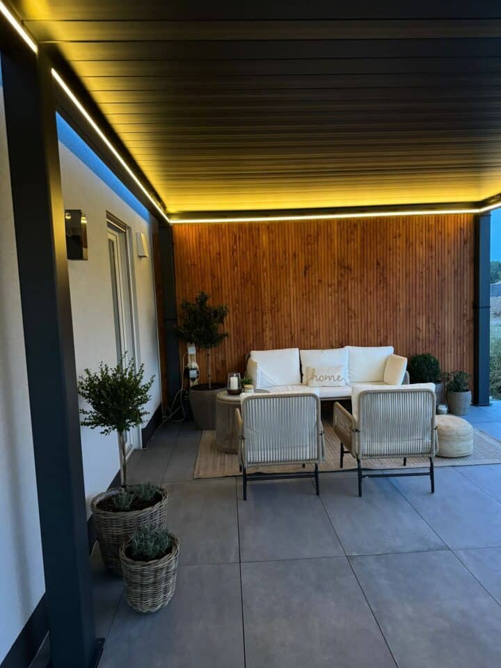 Eine gemütliche Außenterrasse besticht durch ein weißes Sofa mit dekorativem Kissen und zwei gegenüberliegenden Stühlen unter einer eleganten Lamellenüberdachung. Die Decke ist mit warmen Einbauleuchten ausgestattet, und die holzgetäfelte Wand verleiht dem Raum Charme. Topfpflanzen rund um den Fliesenboden sorgen für eine entspannende Atmosphäre.