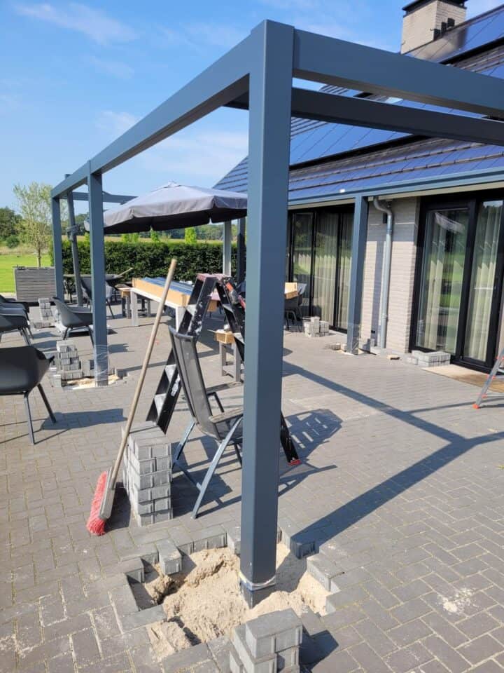 Auf einer Terrasse aus grauen Ziegeln und Sand wird eine moderne Pergola-Struktur für den Außenbereich errichtet. Die Szene, die mit Werkzeugen wie Leitern und einem Besen übersät ist, deutet auf eine nachhaltige Aluminiumüberdachung hin. Im Hintergrund vervollständigen Stühle und Tische mit Sonnenschirmen diese einladende Gaanderen-Kulisse.