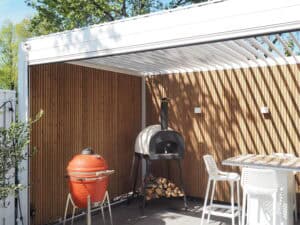 Außenterrasse mit eleganter Lamellenüberdachung und Bambuswand. Verfügt über einen roten Keramikgrill, einen Holzofen mit Holzscheiten darunter und einen weißen Bartisch mit Hockern. Sonnenlicht wirft gestreifte Schatten über den Bereich und schafft ein einladendes Ambiente, das perfekt für jede Kundengeschichte ist.