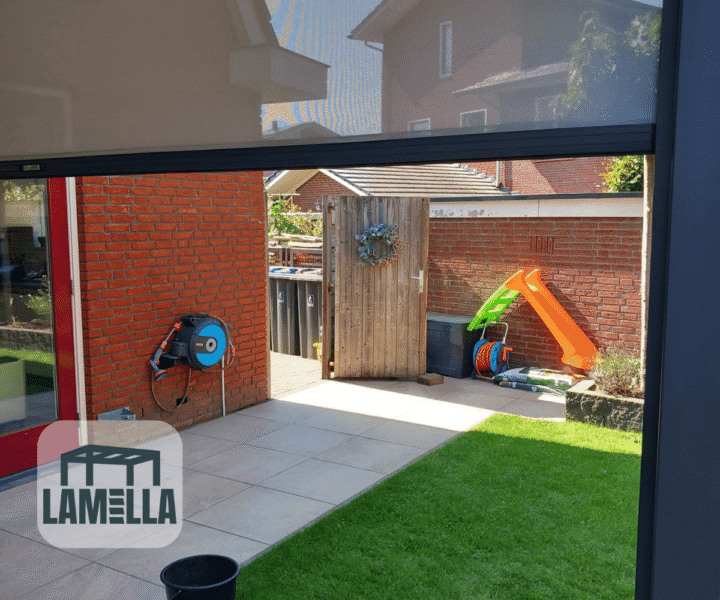 Ein Hinterhof mit Terrasse und grünem Rasen. In der Nähe einer Backsteinmauer befinden sich eine kleine Rutsche und Spielzeug. Links an der Mauer ist ein Gartenschlauch befestigt. Eine Holztür führt zu einem weiteren Bereich. Das „LAMELLA“-Logo befindet sich in der unteren linken Ecke.